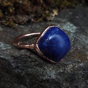 Può includere: Un anello in rame con una pietra preziosa di lapislazzuli blu scuro incastonata in una lunetta quadrata. L'anello ha un aspetto rustico e artigianale.
