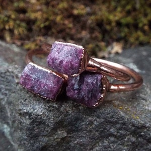 May include: Three copper rings with raw pink ruby stones set in the metal. The rings are stacked on top of each other.