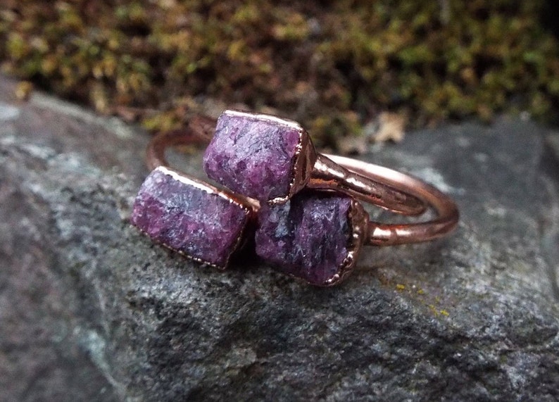 May include: Three copper rings with raw pink ruby stones set in the metal. The rings are stacked on top of each other.