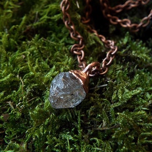 May include: A clear quartz crystal pendant on a copper chain. The pendant is a raw, unpolished crystal with a natural shape. The chain is a simple, linked chain. The pendant is hanging on a bed of green moss.