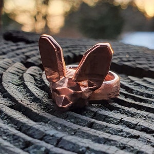 Könnte beinhalten: Ein kupferfarbener Ring mit einem geometrischen Fuchsdesign. Der Ring liegt auf einer dunklen Holzoberfläche.