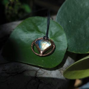 May include: A handcrafted pendant necklace featuring a labradorite gemstone with blue, yellow, and green iridescence. The stone is set in a copper-colored metal frame and hangs from a black cord. The pendant rests on a large, green leaf.