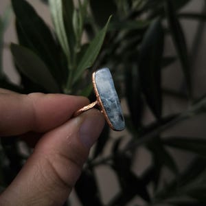 Könnte beinhalten: Ein kupferfarbener Ring mit einem langen, rechteckigen, hellblauen Edelstein. Der Edelstein hat ein leicht durchscheinendes Aussehen. Der Ring wird vor einem Hintergrund aus grünem Laub gehalten.