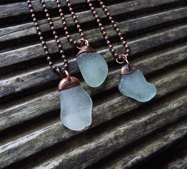 Puede incluir: Tres collares con cadenas de cobre y colgantes de vidrio de mar. El vidrio de mar es de color azul verdoso claro y el cobre es de color marr&oacute;n c&aacute;lido.