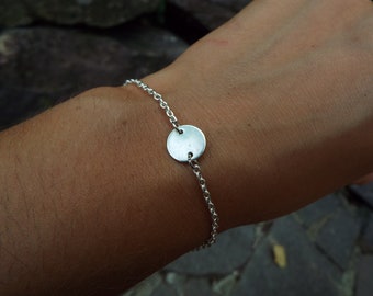 May include: A silver chain bracelet with a round silver charm. The charm is smooth and has a slightly hammered texture.