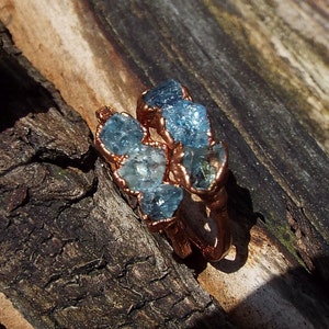 Könnte beinhalten: Ein Ring aus Kupferdraht mit mehreren blauen Aquamarin-Edelsteinen, die in den Draht gefasst sind. Der Ring liegt auf einem Stück verwittertem Holz.