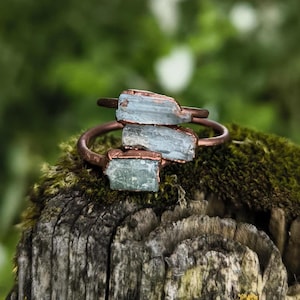 Könnte beinhalten: Drei rohe Aquamarin-Kristallringe, die in Kupferdraht gefasst sind. Die Ringe sind übereinander gestapelt und liegen auf einem Stück Holz mit Moos.