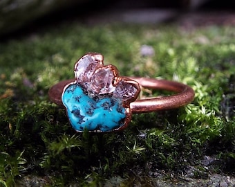 May include: A copper ring featuring a turquoise and clear quartz stone setting. The ring is resting on a bed of green moss.