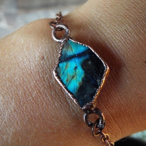 Könnte beinhalten: Ein kupferfarbenes Kettenarmband mit einem rautenförmigen Labradoritstein. Der Stein zeigt schillernde blaue und grüne Farbtöne, eingefasst in eine Kupferfassung. Das Armband ist mit einer filigranen Kette versehen.