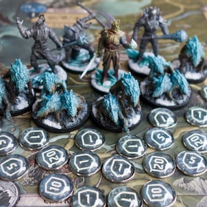 Op de afbeelding: Een bordspelset met beschilderde miniatuurfiguren van personages en wezens. De figuren zijn gerangschikt op een speelbord met tokens en dobbelstenen. De figuren zijn geschilderd in tinten grijs, blauw en wit. De tokens zijn zilverkleurig met zwarte nummers.