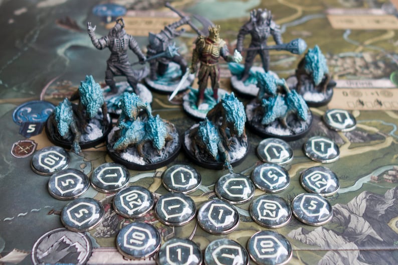 Op de afbeelding: Een bordspelset met beschilderde miniatuurfiguren van personages en wezens. De figuren zijn gerangschikt op een speelbord met tokens en dobbelstenen. De figuren zijn geschilderd in tinten grijs, blauw en wit. De tokens zijn zilverkleurig met zwarte nummers.
