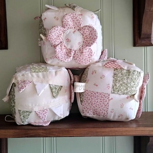 Może przedstawiać: Trzy ręcznie robione kostki z tkaniny z kwiatowymi i patchworkowymi wzorami. Jedna kostka ma duży różowy kwiat, druga girlandę z małych trójkątów, a trzecia wzór patchworkowy. Są one wystawione na drewnianej półce na tle jasnozielonej ściany.