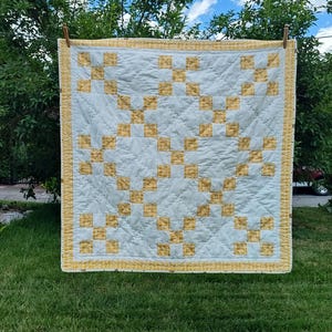May include: A square quilt featuring a repeating pattern of light blue fabric squares interspersed with yellow and white checkered fabric squares. The quilt is finished with a yellow and white checkered border and has visible quilted stitching.