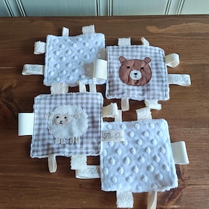 Pode incluir: Um brinquedo sensorial para bebês com quatro peças de tecido quadradas. Dois quadrados são brancos com um padrão de bolinhas, e dois são xadrez com um desenho de urso e ovelha. Cada quadrado tem uma etiqueta de fita e uma etiqueta.