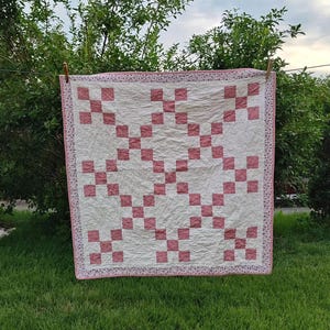 Può includere: Una trapunta quadrata con un motivo ripetitivo di quadrati color rosa antico disposti a griglia su uno sfondo bianco sporco testurizzato. La trapunta è rifinita con un bordo sottile caratterizzato da una piccola stampa floreale rosa e bianca.
