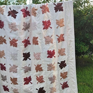 May include: A white quilt featuring a repeating pattern of appliquéd fabric leaves in various shades of red, brown, orange, and tan. The leaves are arranged in vertical columns across the surface, showcasing diverse patterns like gingham, floral, and solid textures.
