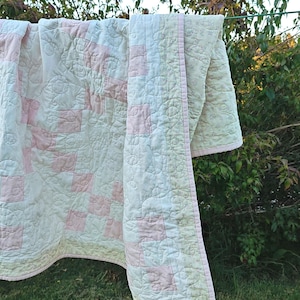 May include: A quilted blanket featuring a patchwork pattern of soft pink and cream-colored squares. The fabric displays an intricate, swirling stitched design throughout, finished with a light pink border and a subtle floral print on the reverse side.