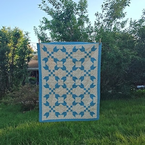 May include: A rectangular quilt featuring a repeating geometric pattern of light blue star-like shapes on a cream-colored background. The quilt is finished with a solid light blue border and exhibits visible stitching throughout the fabric surface.
