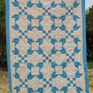 May include: A handmade quilt with a repeating star pattern in light blue and cream. The quilt is rectangular with a blue border and is hanging outside on a clothesline. The star pattern is made of squares and triangles.