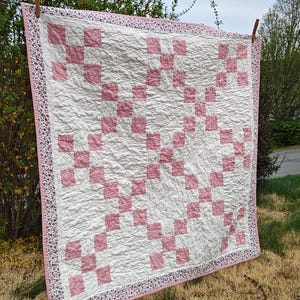 May include: A handmade quilt with a white background and a pink checkerboard pattern. The quilt is hanging on a clothesline outdoors. The border is a pink floral print.