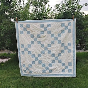 Może przedstawiać: Kwadratowy quilt z powtarzającym się wzorem jasnoniebieskich i białych kwadratów z tkaniny ułożonych w siatkę. Quilt ma jednolity jasnoniebieski brzeg i jest wykończony widocznymi przeszyciami na całej powierzchni, co nadaje mu teksturę.