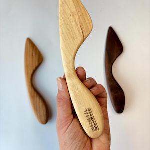 May include: Three wooden butter spreaders in varying shades of brown. The central spreader is held in a hand, displaying a light wood grain. The handle is curved, and the blade is triangular. The text "Hand Crafted by MfamWood" is visible.
