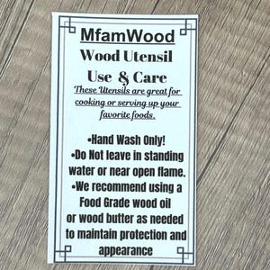 Puede incluir: Una tarjeta rectangular blanca con texto negro sobre una superficie de madera. La tarjeta dice "MfamWood Wood Utensil Use & Care" y proporciona instrucciones de cuidado para utensilios de madera, incluyendo lavado a mano y recomendaciones de aceite.