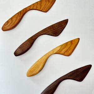 Puede incluir: Cuatro esparcidores de madera dispuestos en diagonal, cada uno con un mango curvo y una hoja plana y angulada. El conjunto incluye dos esparcidores de color marr&oacute;n oscuro, uno marr&oacute;n medio con veta visible y uno de color tostado claro.