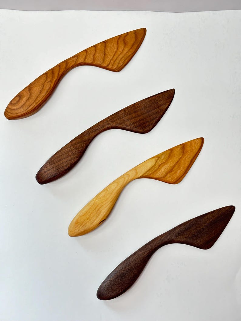 Puede incluir: Cuatro esparcidores de madera dispuestos en diagonal, cada uno con un mango curvo y una hoja plana y angulada. El conjunto incluye dos esparcidores de color marr&oacute;n oscuro, uno marr&oacute;n medio con veta visible y uno de color tostado claro.