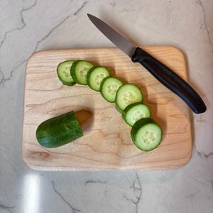May include: A wooden cutting board with a partially sliced cucumber and a black-handled knife. The cucumber is cut into green slices, with the remaining portion of the cucumber on the board. The knife is resting on the board.