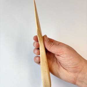 Puede incluir: Un abrecartas de madera de color claro, liso, con una punta afilada y un mango redondeado y ergon&oacute;mico. La veta natural de la madera es visible en toda la superficie, mostrando un acabado pulido y una silueta esbelta y alargada.