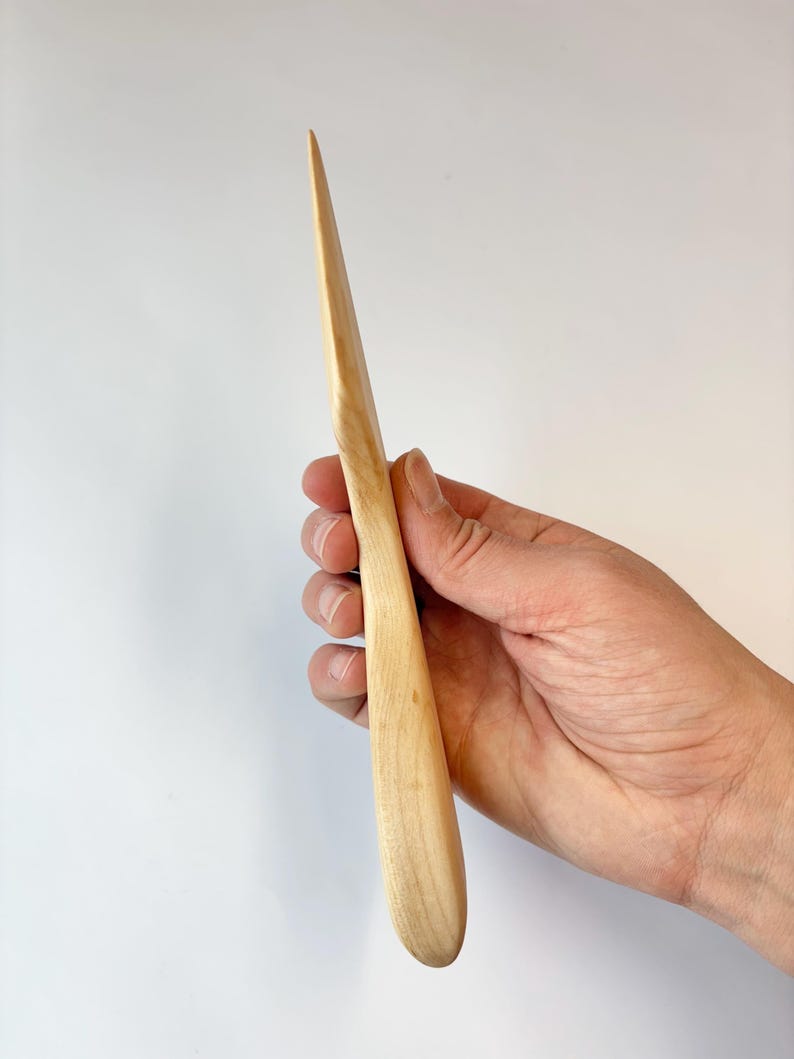 Puede incluir: Un abrecartas de madera de color claro, liso, con una punta afilada y un mango redondeado y ergon&oacute;mico. La veta natural de la madera es visible en toda la superficie, mostrando un acabado pulido y una silueta esbelta y alargada.