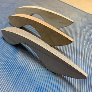 May include: Three curved wooden handles arranged in a diagonal row. The top handle is light beige, the middle handle is a medium tan, and the bottom handle is a dark gray. Each handle features a smooth, elongated shape with a tapered end.