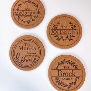 May include: Four round cork coasters with engraved family names and decorative leaf designs. The coasters feature the names "McCormick family", "The Johnsons", "The Monks", and "The Brock Family". The Monks coaster also includes the text "It's so good to be home".
