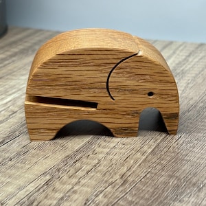May include: A wooden elephant figurine with a curved trunk and small eye. The elephant is made of light brown wood with visible grain. The figurine has a slot on the side, suggesting it may be a phone holder or a decorative item.