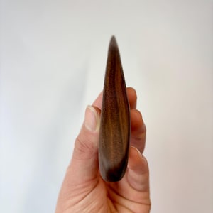 Puede incluir: Una herramienta de madera de color marr&oacute;n oscuro, lisa, con un extremo puntiagudo y una base redondeada. El objeto presenta un patr&oacute;n de veta de madera visible con varios tonos de marr&oacute;n, mostrando un acabado pulido y una forma curva.