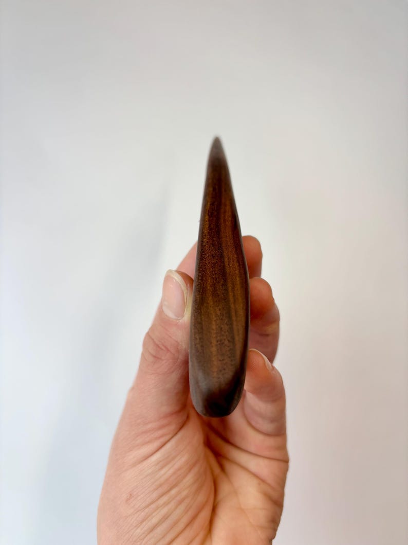 Puede incluir: Una herramienta de madera de color marr&oacute;n oscuro, lisa, con un extremo puntiagudo y una base redondeada. El objeto presenta un patr&oacute;n de veta de madera visible con varios tonos de marr&oacute;n, mostrando un acabado pulido y una forma curva.