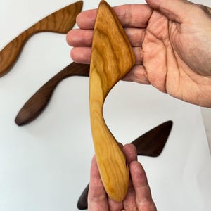 Puede incluir: Un esparcidor de madera de color claro con un mango curvo y una hoja plana e inclinada. La herramienta presenta un patr&oacute;n de veta de madera natural visible. Otros esparcidores en tonos de madera m&aacute;s oscuros se sit&uacute;an al fondo sobre una superficie blanca.