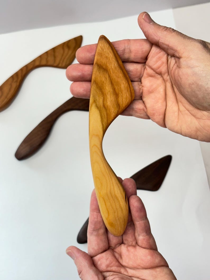 Puede incluir: Un esparcidor de madera de color claro con un mango curvo y una hoja plana e inclinada. La herramienta presenta un patr&oacute;n de veta de madera natural visible. Otros esparcidores en tonos de madera m&aacute;s oscuros se sit&uacute;an al fondo sobre una superficie blanca.