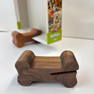 May include: A wooden dog bone-shaped phone stand in a medium brown color. The stand has a slot for holding a phone and is designed to prop up a phone. The stand is made of wood and has a smooth finish.