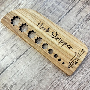 May include: Wooden herb stripper with flower-shaped cutouts and the words "Herb Stripper" engraved on the surface. A decorative floral design is etched on the right side. The tool is made of light-colored wood.