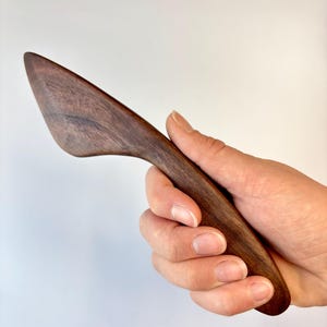 Puede incluir: Un esparcidor de madera de color marr&oacute;n oscuro con una hoja curva y c&oacute;nica, y un acabado suave y pulido. El utensilio presenta un patr&oacute;n de veta de madera distintivo en toda su superficie, mostrando un color marr&oacute;n natural profundo con variaciones.