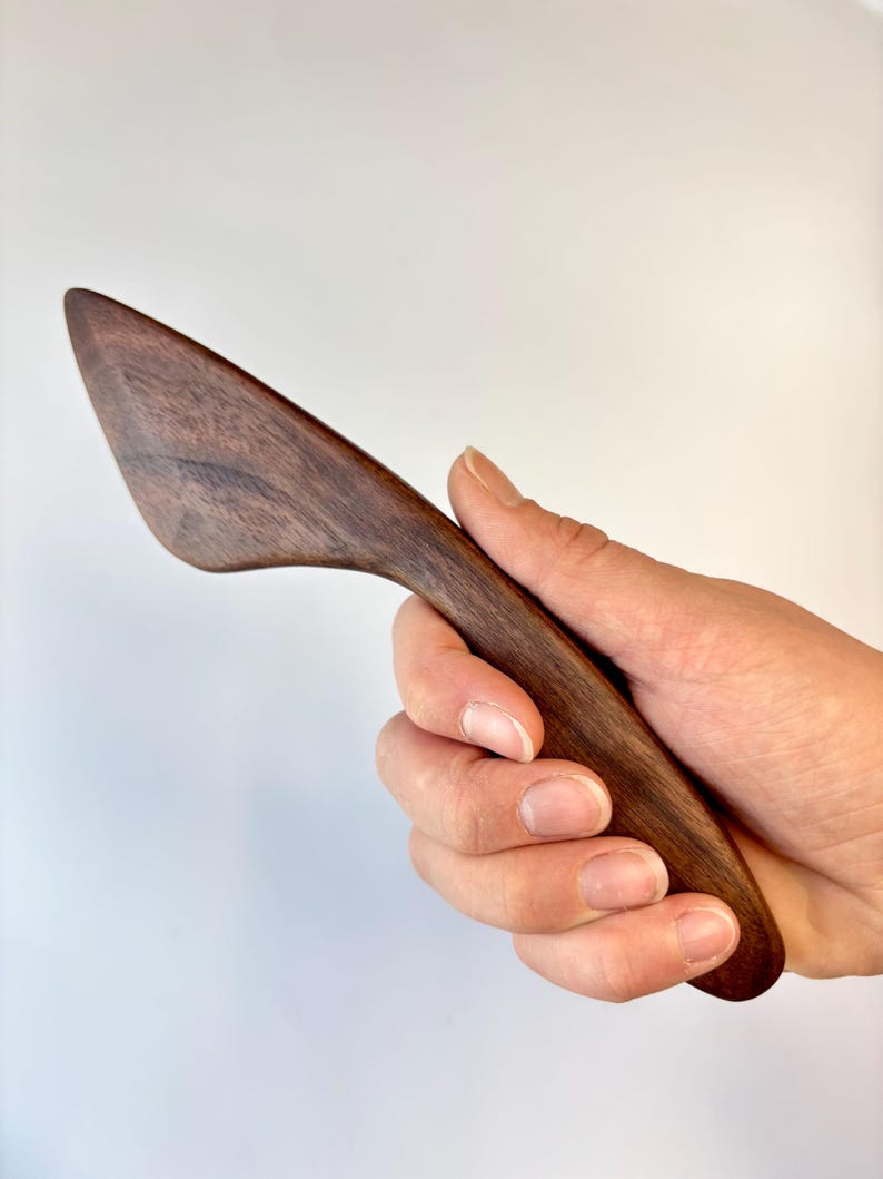 Puede incluir: Un esparcidor de madera de color marr&oacute;n oscuro con una hoja curva y c&oacute;nica, y un acabado suave y pulido. El utensilio presenta un patr&oacute;n de veta de madera distintivo en toda su superficie, mostrando un color marr&oacute;n natural profundo con variaciones.