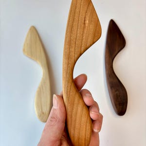 Puede incluir: Tres esp&aacute;tulas de cocina de madera con mangos curvos y hojas puntiagudas. Una esp&aacute;tula es de color beige claro, otra es de color marr&oacute;n medio con vetas de madera visibles y la tercera es de color marr&oacute;n oscuro. Cada herramienta tiene una forma ergon&oacute;mica.
