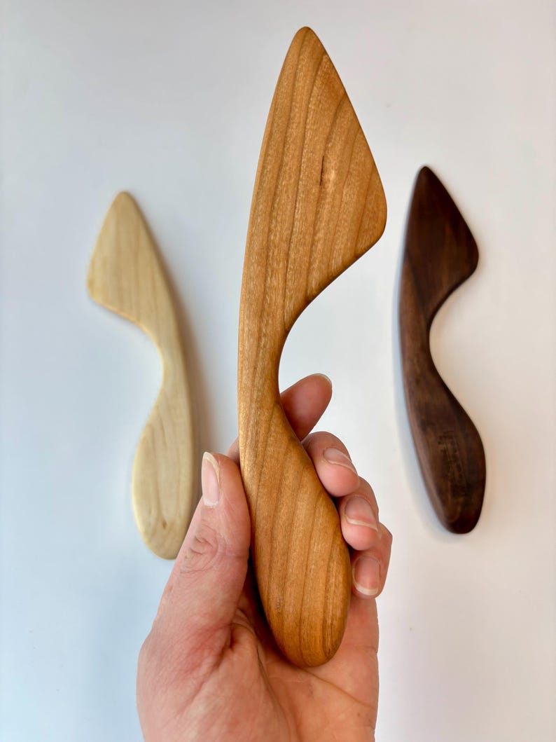 Puede incluir: Tres esp&aacute;tulas de cocina de madera con mangos curvos y hojas puntiagudas. Una esp&aacute;tula es de color beige claro, otra es de color marr&oacute;n medio con vetas de madera visibles y la tercera es de color marr&oacute;n oscuro. Cada herramienta tiene una forma ergon&oacute;mica.