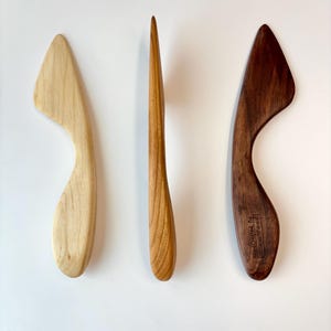 Puede incluir: Tres herramientas de madera con formas suaves y curvas. La herramienta de la izquierda es de color beige claro, la central es una pieza delgada de color marr&oacute;n medio y la de la derecha es marr&oacute;n oscuro con un sello Hand Crafted by Seven Mile Wood.