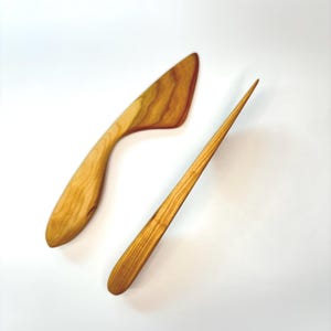 Puede incluir: Dos utensilios de cocina hechos de madera de color marr&oacute;n claro con vetas visibles. Uno presenta una forma de hoja triangular ancha y curva, mientras que el otro es una herramienta larga, delgada y puntiaguda con mango redondeado y punta c&oacute;nica.