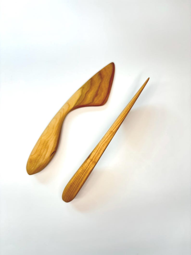 Puede incluir: Dos utensilios de cocina hechos de madera de color marr&oacute;n claro con vetas visibles. Uno presenta una forma de hoja triangular ancha y curva, mientras que el otro es una herramienta larga, delgada y puntiaguda con mango redondeado y punta c&oacute;nica.