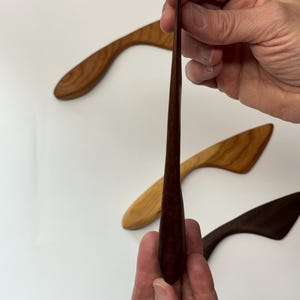 Puede incluir: Un conjunto de tres esparcidores de madera con mangos curvos y hojas c&oacute;nicas. Un esparcidor de color marr&oacute;n oscuro se sostiene verticalmente en primer plano, mientras que otros dos, uno marr&oacute;n claro y otro oscuro, se sit&uacute;an detr&aacute;s.