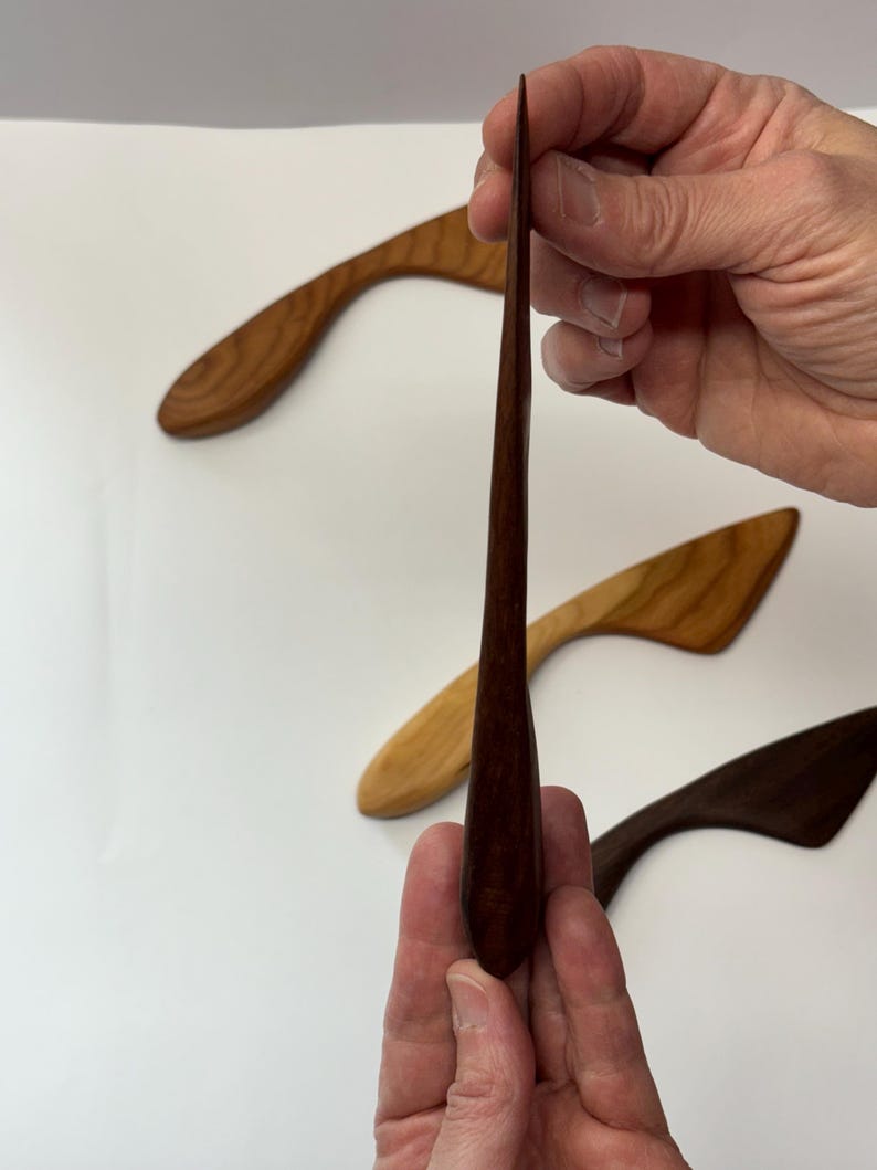 Puede incluir: Un conjunto de tres esparcidores de madera con mangos curvos y hojas c&oacute;nicas. Un esparcidor de color marr&oacute;n oscuro se sostiene verticalmente en primer plano, mientras que otros dos, uno marr&oacute;n claro y otro oscuro, se sit&uacute;an detr&aacute;s.